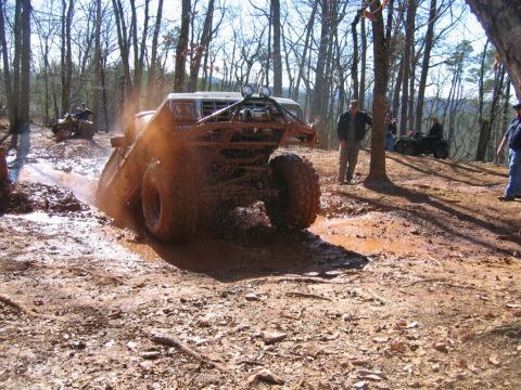 1992 Ford Mustang - An off-road vehicle navigates a muddy trail, kicking up water and dirt. Other ATVs are visible in the background.