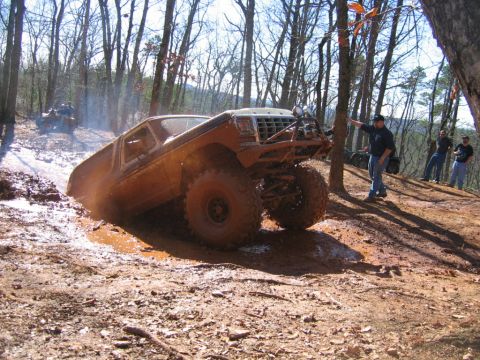 1992 Ford Mustang - A heavily modified off-road truck navigates a muddy trail with spectators observing the background.