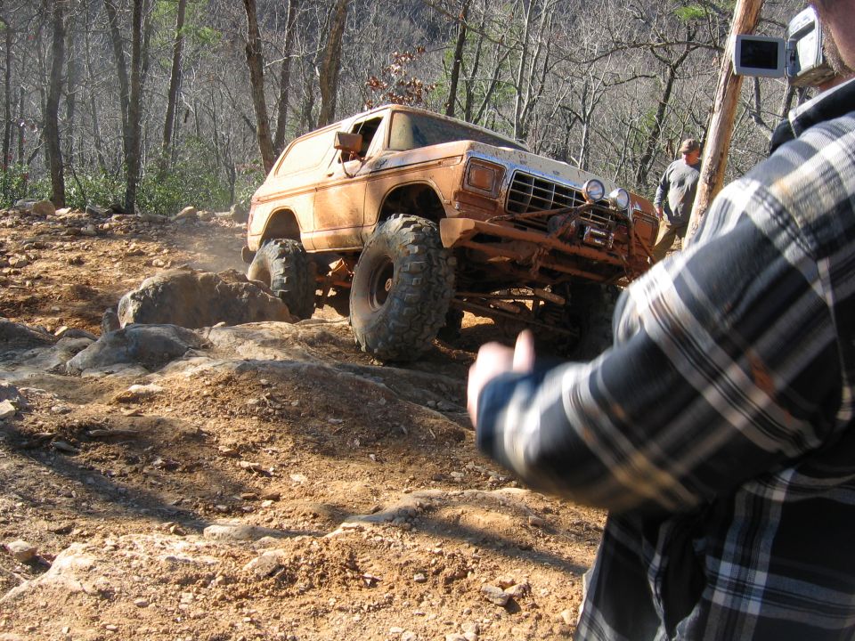 Orange off-road SUV with large tires climbing a rocky hill. Person in plaid shirt filming