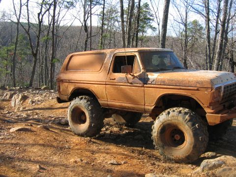 1992 Ford Mustang - This lifted SUV is covered in mud and parked on a rocky, off-road trail surrounded by trees.