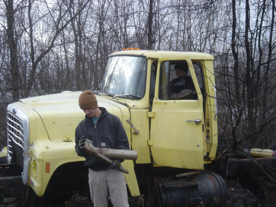 Guy holding exhaust pipe next to yellow semi-truck cab. Another person sits in the driver's seat