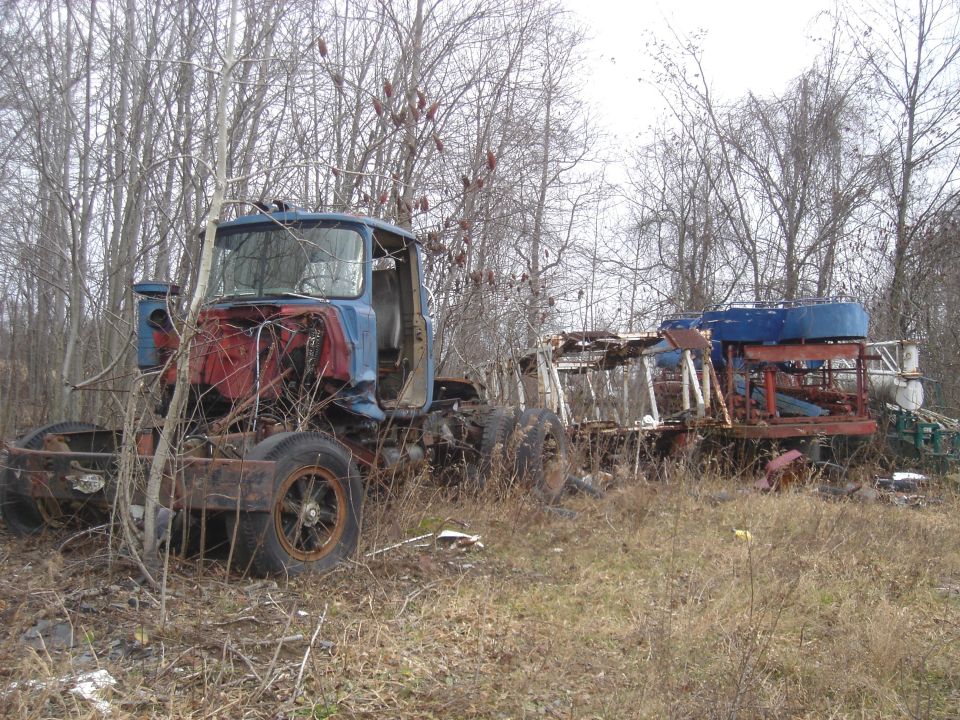 Blue and red truck cab with missing grille, next to a rusty trailer with blue tanks, in a dry, overgrown field