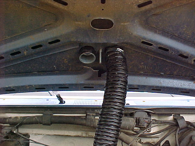 Underhood view showing a flexible black duct hose attached to a silver pipe, with visible wiring and part of the windshield wipers