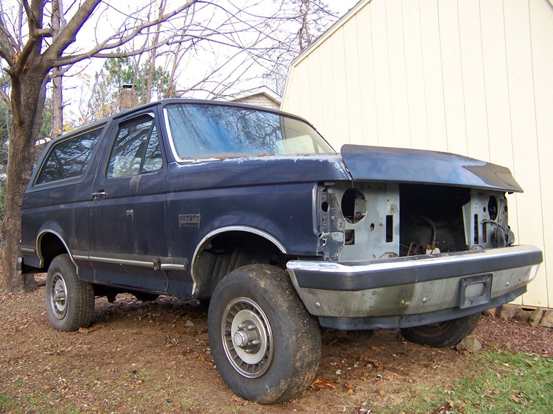 Dark blue 1987 Ford Bronco with missing headlights and grille, parked on dirt