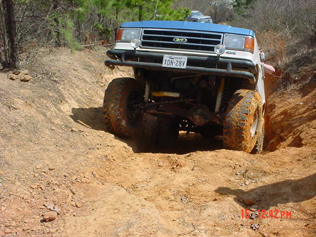 Blue Ford 4x4 climbing out of a red-dirt rut on a pine trail, muddy tires and Texas plate XDN-28V