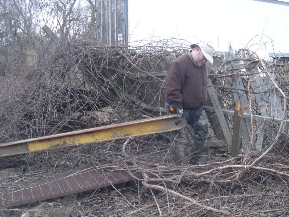 Man in a brown jacket and camouflage pants carries a yellow metal beam through a junkyard filled with branches and debris