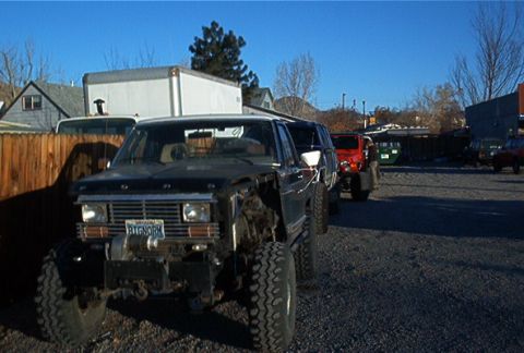 1986 Ford Bronco - A damaged black 1987 Ford Bronco with off-road tires is parked in a lot with other vehicles under a clear blue sky.