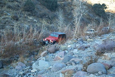 1986 Ford Bronco - A red Jeep Wrangler navigates a rough, rocky terrain, with other vehicles visible further down the path.