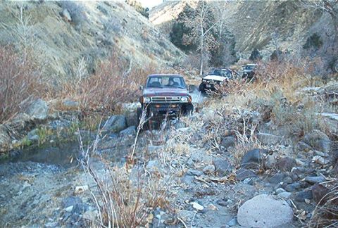 1986 Ford Bronco - A red SUV navigates a challenging off-road path, with other vehicles following in the distance.