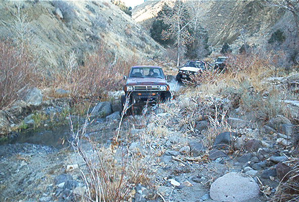 Red SUV driving on a rocky, dry creek bed with other vehicles behind it