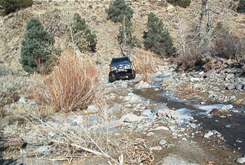 1986 Ford Bronco - A dark blue SUV is shown off-roading through a rocky stream bed in a dry, hilly landscape.