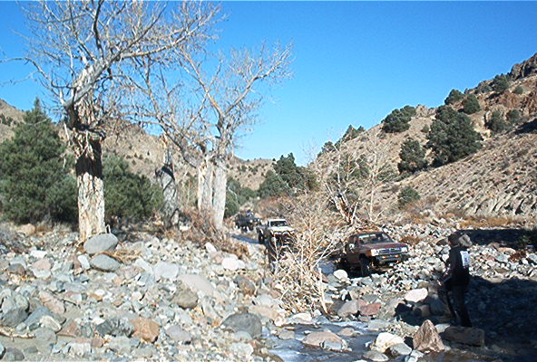 Several off-road vehicles navigate a rocky, dry riverbed with sparse trees and hills under a clear blue sky