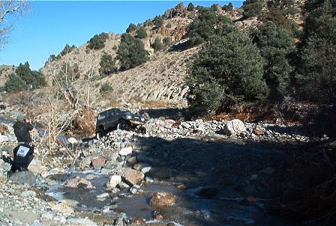 1986 Ford Bronco - A dark SUV attempts to traverse a difficult, rocky stream bed under a clear blue sky.