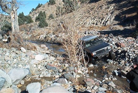 1986 Ford Bronco - A black SUV is shown off-roading through a rocky stream bed in a dry, mountainous landscape.