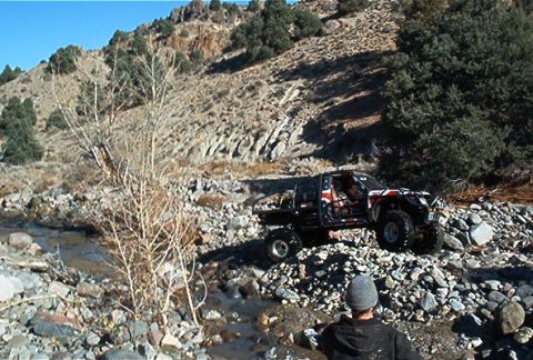 1986 Ford Bronco - Custom painted off-road truck navigates a rocky stream bed on a sunny day.