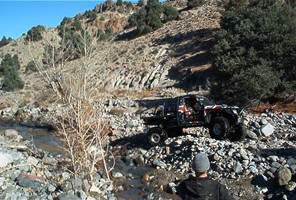 Off-road truck with custom paint driving over rocks near a stream, person in foreground