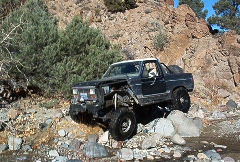 1986 Ford Bronco - This modified SUV is shown off-roading on a rocky trail with sparse vegetation and a steep hillside.