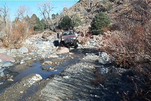 1986 Ford Bronco - A red SUV drives through a rocky stream bed in a dry, mountainous area.