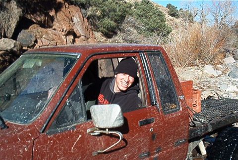 1986 Ford Bronco - Driver smiles from inside a mud-splattered red pickup truck with a flatbed in the background.