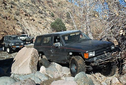 1986 Ford Bronco - Multiple off-road vehicles traverse a challenging, rocky trail in a dry, mountainous area.