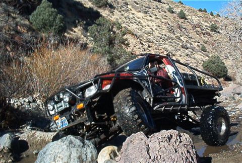 1986 Ford Bronco - Modified off-road vehicle with a prominent roll cage traverses a rocky stream bed.