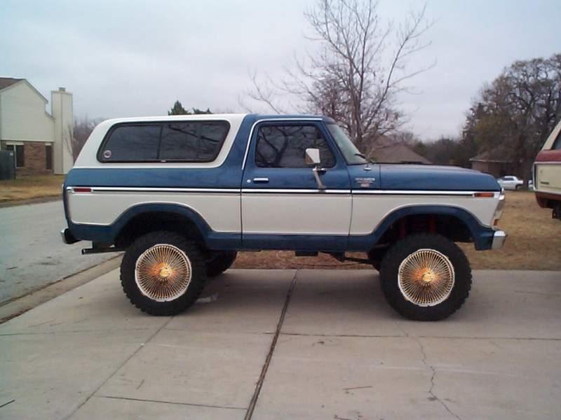 Blue and white Ford Bronco with gold wire wheels, lifted suspension, parked on concrete