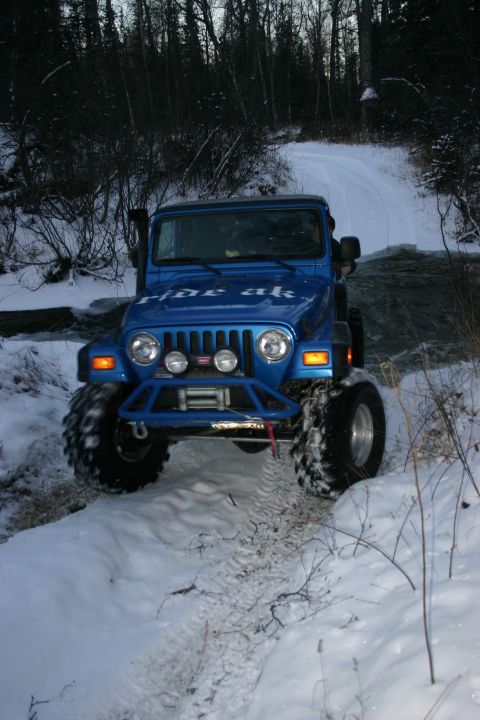 2003 Jeep TJ - A blue Jeep Wrangler navigates a snowy trail, with "ride ak" visible on its hood.