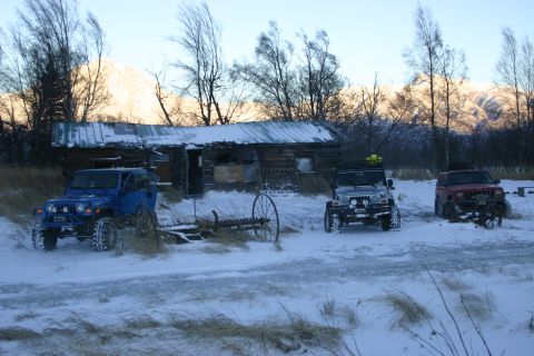 2003 Jeep TJ - Three Jeeps are parked in the snow outside a weathered wooden cabin, with snow-covered mountains visible behind the trees.