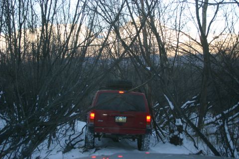 2003 Jeep TJ - Rear view of a red Jeep Cherokee on a snow-covered trail surrounded by bare trees.