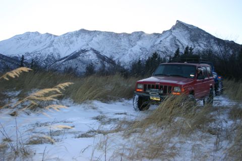 2003 Jeep TJ - A red Jeep Cherokee equipped with a winch is parked in a snowy, grassy area with mountains in the background. Another vehicle is visible be…