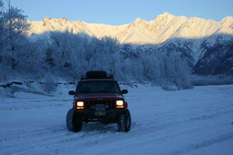 2003 Jeep TJ - A red Jeep Cherokee XJ equipped with a winch and roof rack navigates a snowy landscape with snow-covered mountains in the background.
