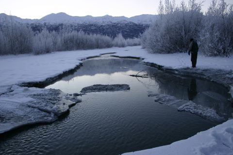 2003 Jeep TJ - A lone figure observes a partially frozen river in a snow-covered landscape with mountains in the distance.