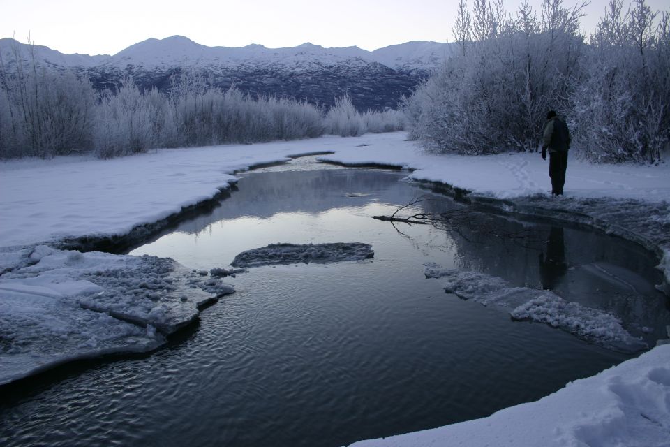 Person standing by a partially frozen river with snow-covered trees and mountains in the background