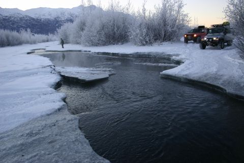 2003 Jeep TJ - Two SUVs navigate a snowy landscape beside a partially frozen river, with snow-covered trees and mountains in the background.