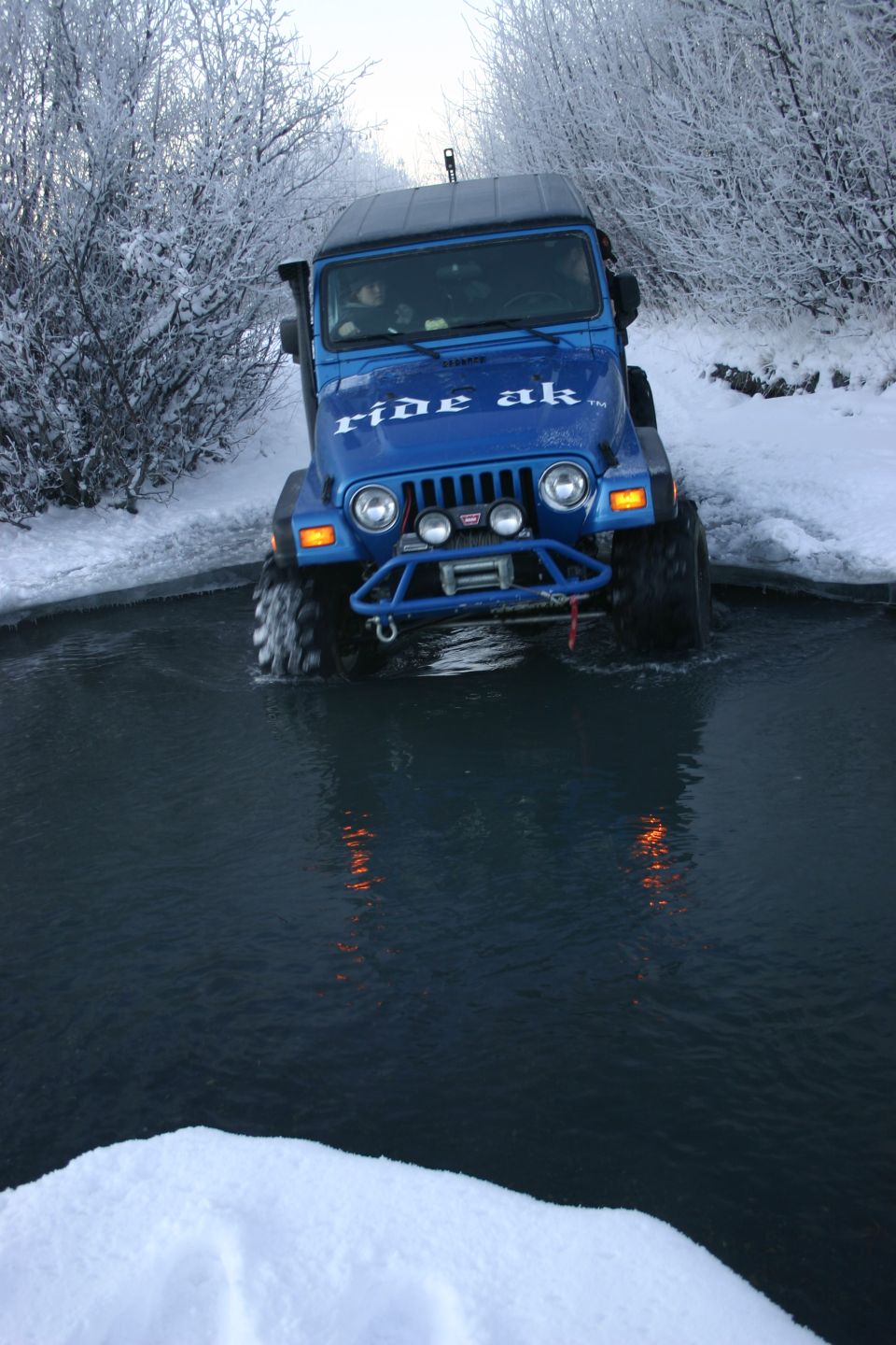 Blue Jeep Wrangler driving through water with snow on the trees. 'ride ak' is on the hood
