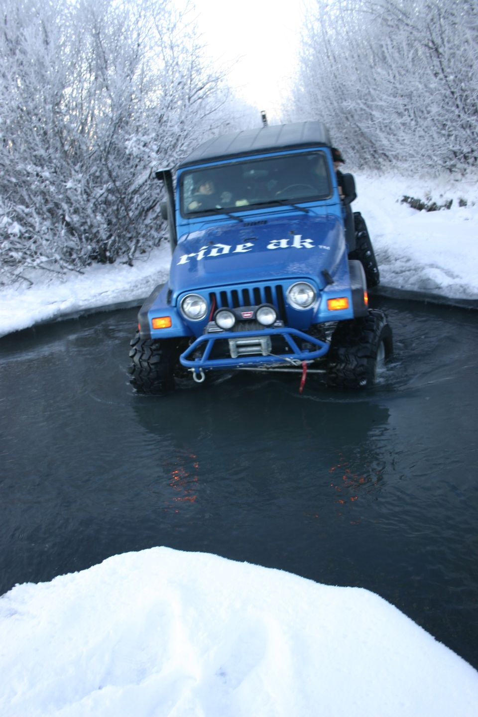 Blue 2003 Jeep TJ driving through water with "ride ak" text on hood