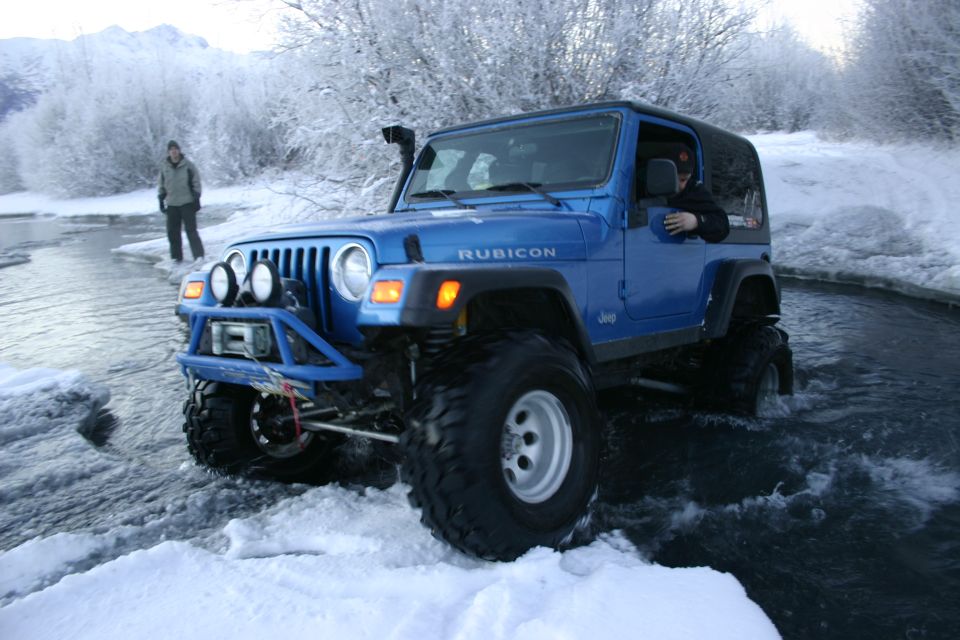 Blue 2003 Jeep TJ Rubicon driving through icy water with snow-covered trees and mountains in background