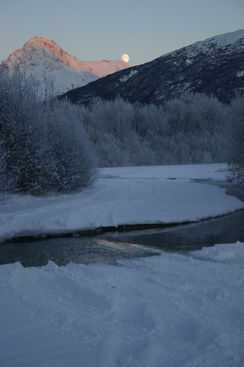 Moon rising over snow-covered mountains and a partially frozen river