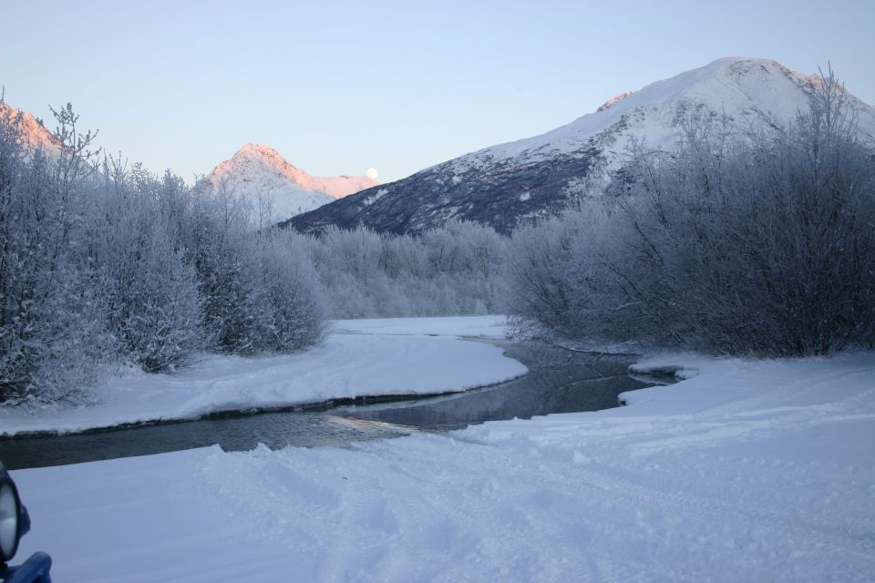Snowy mountains and a river with frost-covered trees under a clear sky. A full moon is visible