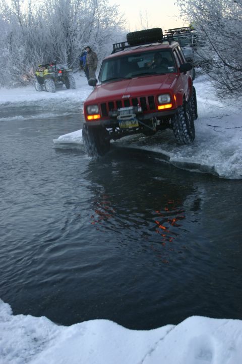 2003 Jeep TJ - A red Jeep Cherokee XJ navigates icy water, with another vehicle and ATVs in the background.