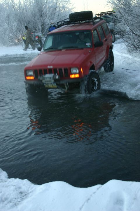2003 Jeep TJ - A red Jeep Cherokee XJ navigates through a shallow body of water in a snowy, wooded area, with another vehicle visible in the background.