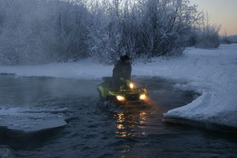 2003 Jeep TJ - An ATV navigates through frigid, icy water with steam rising around it, set against a backdrop of snow-covered trees.