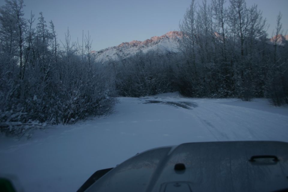 Hood of a vehicle in snow, looking out at a snowy road and frosted trees with a sunlit mountain range in the background