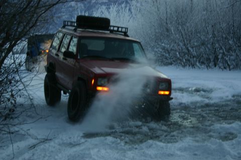 2003 Jeep TJ - A red Jeep Cherokee XJ navigates a snowy path, kicking up snow, with another vehicle visible in the background.