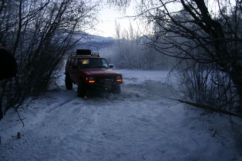 2003 Jeep TJ - A red Jeep Cherokee XJ is stuck in the snow and being pulled by a cable through a wooded area.
