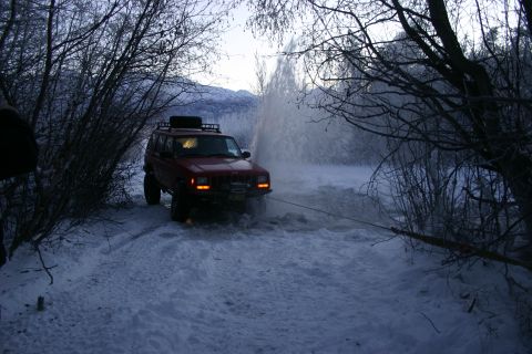 2003 Jeep TJ - A red Jeep Cherokee XJ navigates a snowy terrain, its front tires submerged in slush as a tow rope is attached.