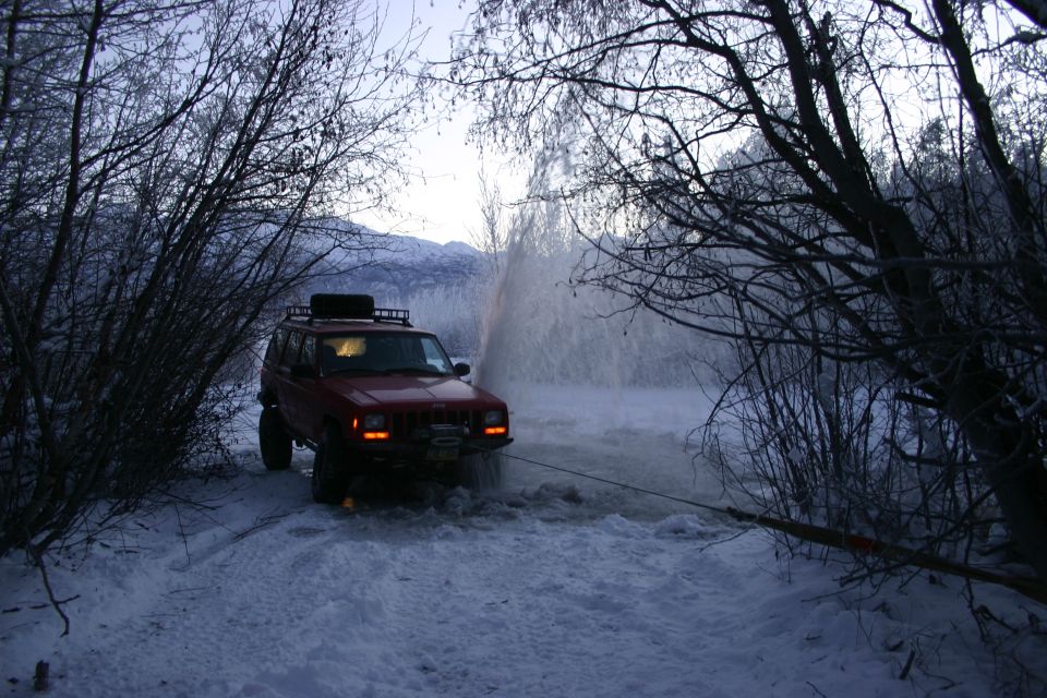 Red Jeep Cherokee XJ stuck in snow, being pulled by a cable. Trees and water spray in background