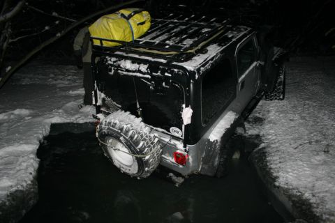 2003 Jeep TJ - A Jeep Wrangler is stuck in a water-filled ditch during a snowy night, with a person visible near the roof rack.