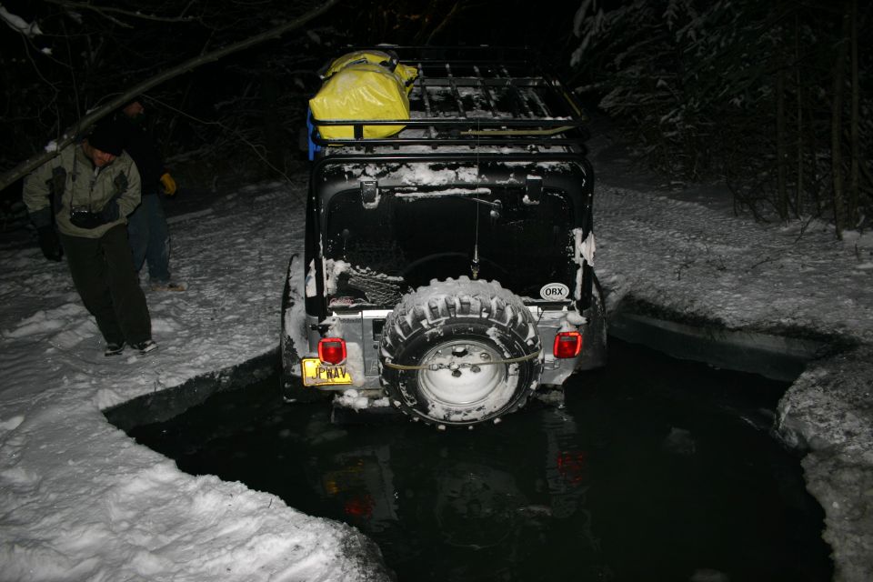 Black 2003 Jeep TJ with a roof rack and yellow bag, partially submerged in dark water with snow on the ground