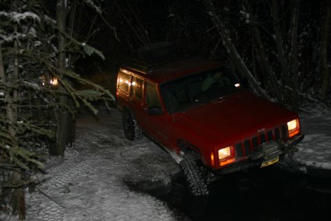 2003 Jeep TJ - Red Jeep Cherokee XJ navigates a snowy path at night, equipped with a winch and spare tire.